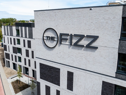 Het gebouw van The Fizz met moderne architectuur en een grote naam op de gevel, omringd door groene beplanting en een stoep.