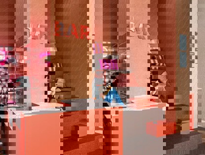 Een moderne bar met een neonbord, een klant achter de balie en een tweede persoon die op de trappen zit met kussens, genietend van een boek.