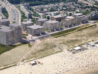 Luchtfoto van een kustgebied met moderne appartementen, brede promenades en een druk strand vol mensen en parasols.