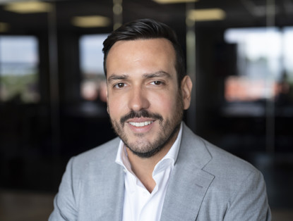 Man in grijze blazer met opgestroopte mouwen, zittend in een modern kantoor met grote ramen op de achtergrond.