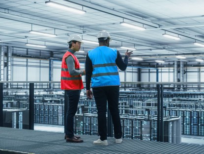 Twee mannen in veiligheidskleding, één met een rood en de ander met een blauw vest, kijken naar een datacenter vol servers.