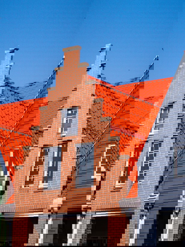 Drie naast elkaar gelegen gebouwen met verschillende gevels; het middelste gebouw heeft een bakstenen muur en een hoog puntdak, omringd door gebouwen met groene en grijze gevels en oranje daken.